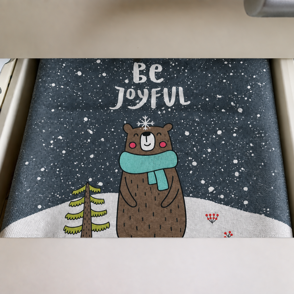 A tea towel laid in a drawer, printed on a slate-grey ground with Be Joyful above a bear in a teal scarf standing in snow