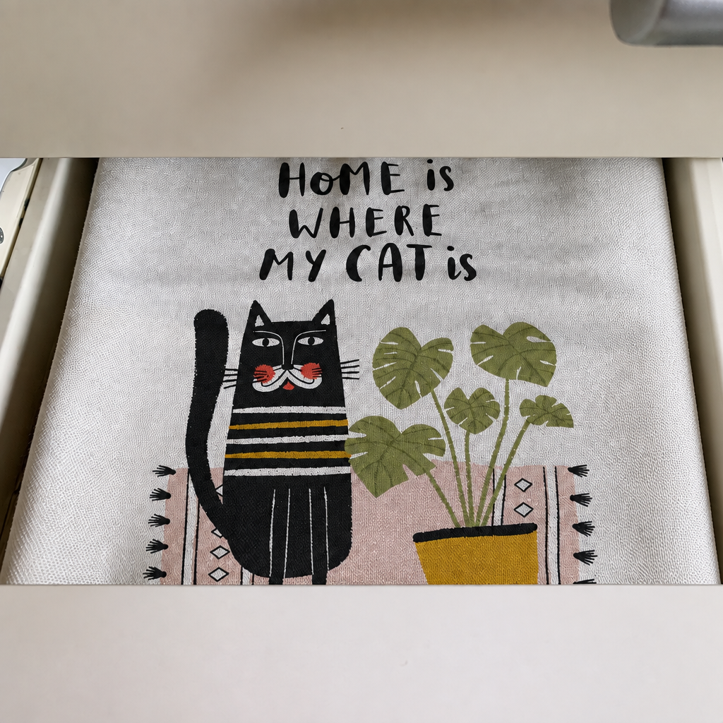 A tea towel laid in a drawer, printed with Home is where my cat is above a striped black cat and a monstera in a yellow pot