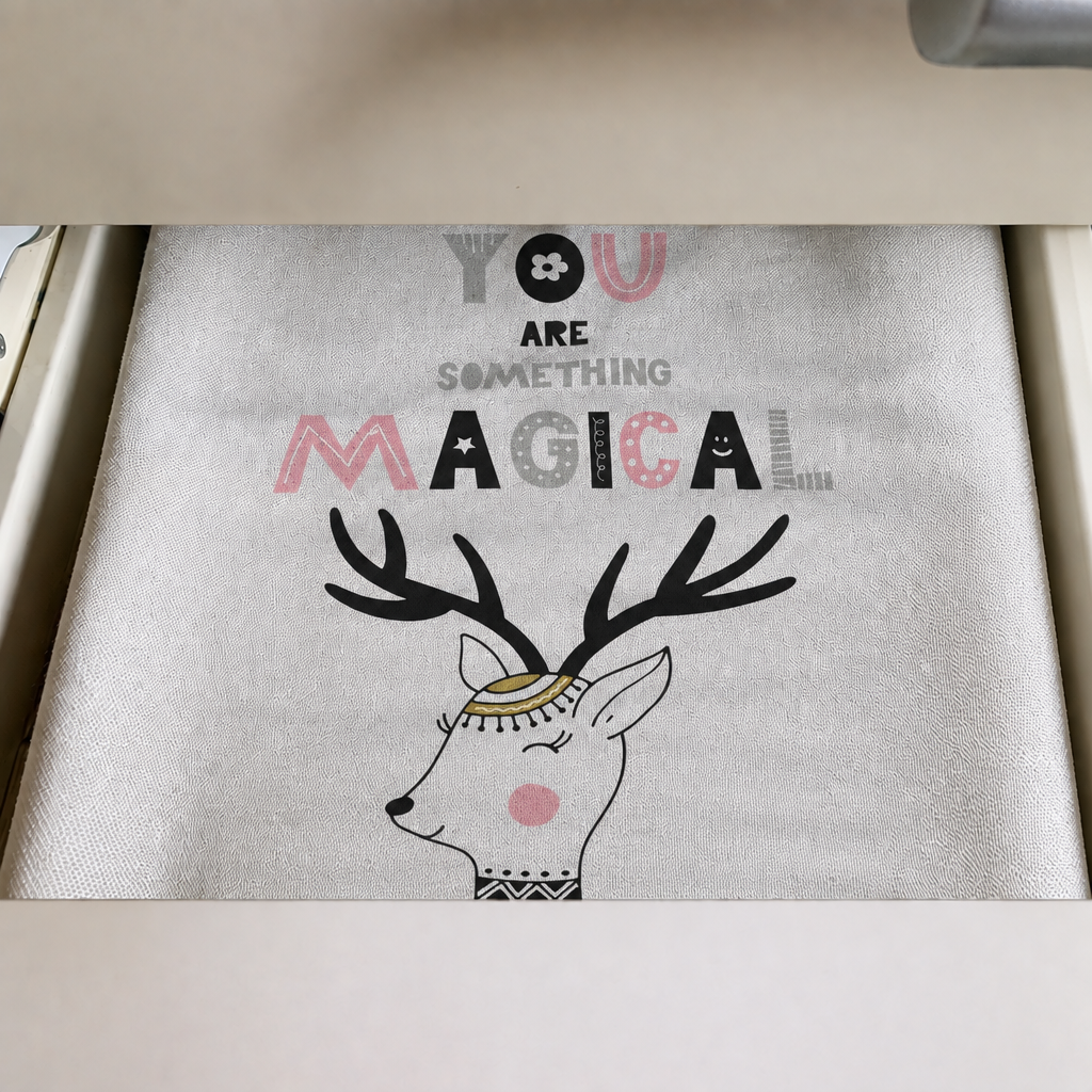 A tea towel laid in a drawer, printed on a pale grey ground with You are something magical above a reindeer in a small gold cap