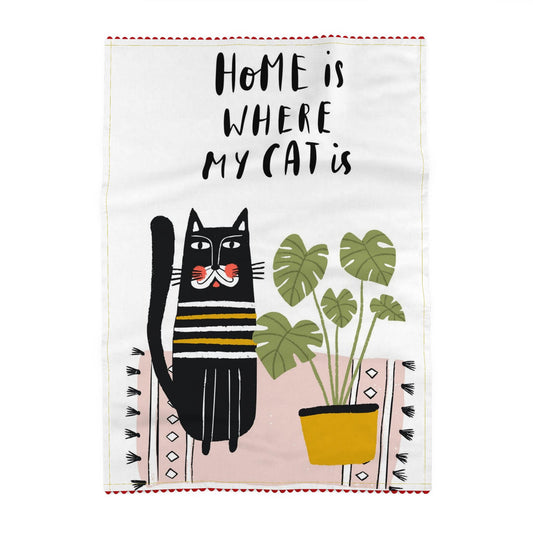 Tea towel reading Home is where my cat is, above a striped black cat and a monstera in a yellow pot on a pink patterned rug with tassels