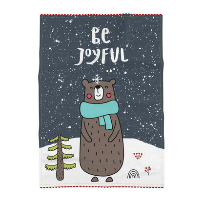 Tea towel printed on a slate-grey ground, reading Be Joyful, above a brown bear in a teal scarf standing in falling snow beside a small fir tree