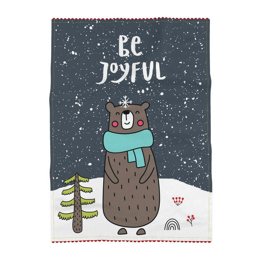 Tea towel printed on a slate-grey ground, reading Be Joyful, above a brown bear in a teal scarf standing in falling snow beside a small fir tree